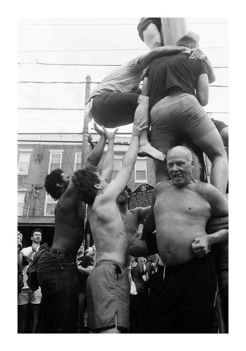 A group of men climb a greased pole in a street.