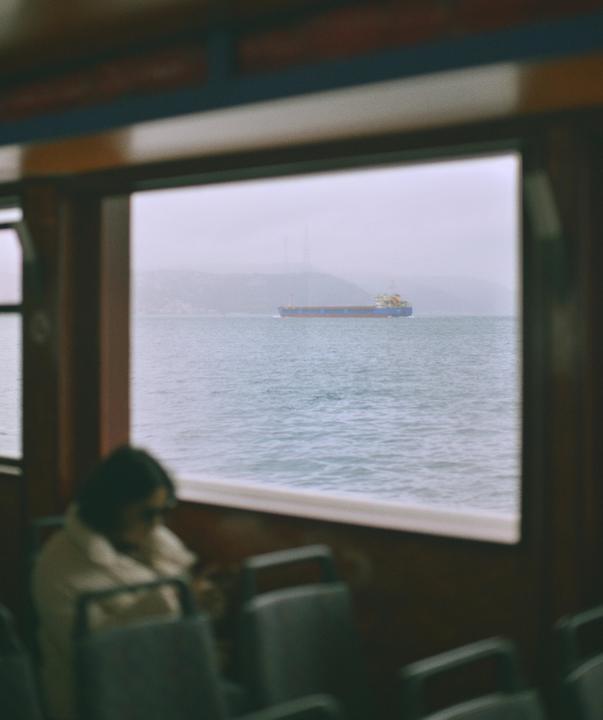A woman sits inside a boat looking out a window at a ship on the water.