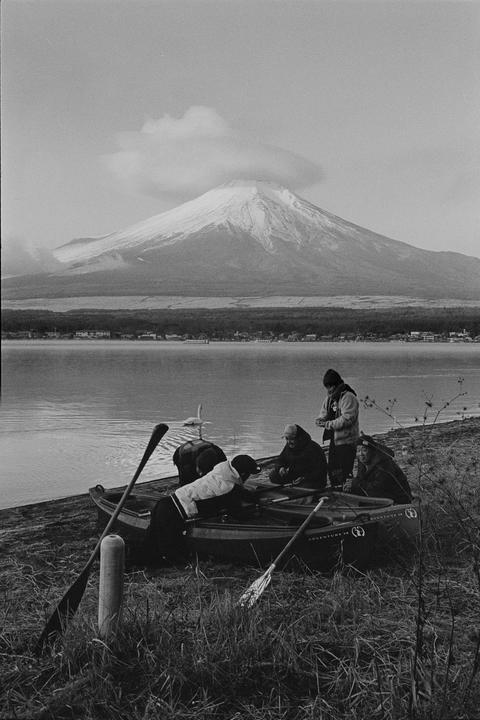 Four people are gathered around a canoe on the shore of Lake Yamanaka, with Mount Fuji in the background.