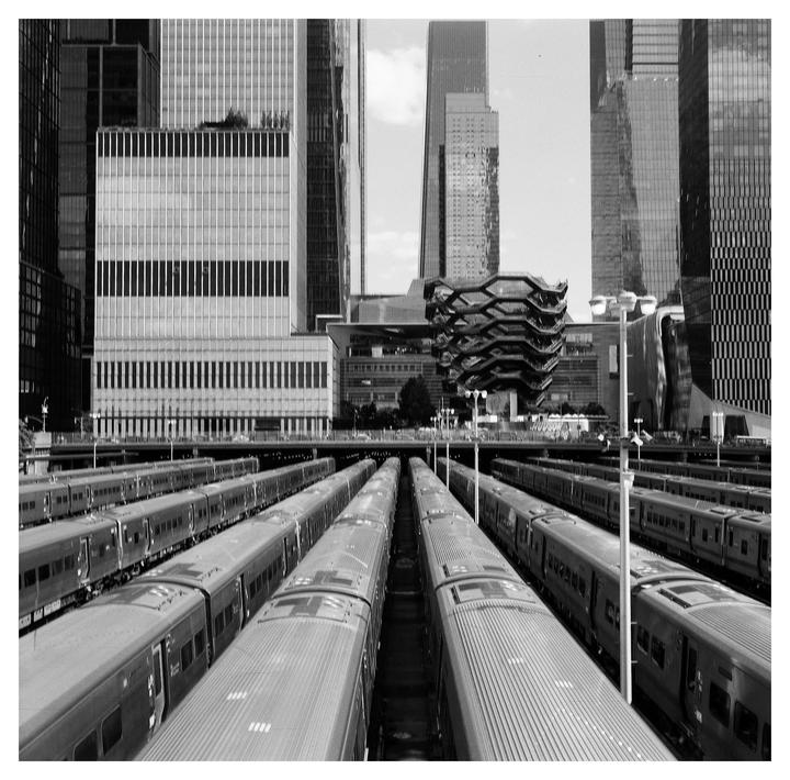 A black and white photograph shows multiple trains parked in a railyard with tall buildings in the background.
