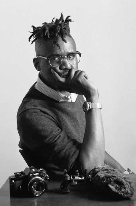 A man with dreadlocks and glasses sits at a table with a camera and microphone.