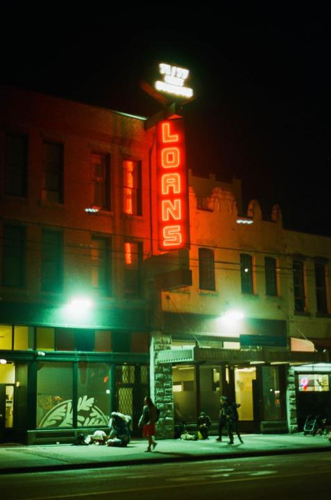 A red neon sign for "LOANS" illuminates a street at night, with people on the sidewalk below.