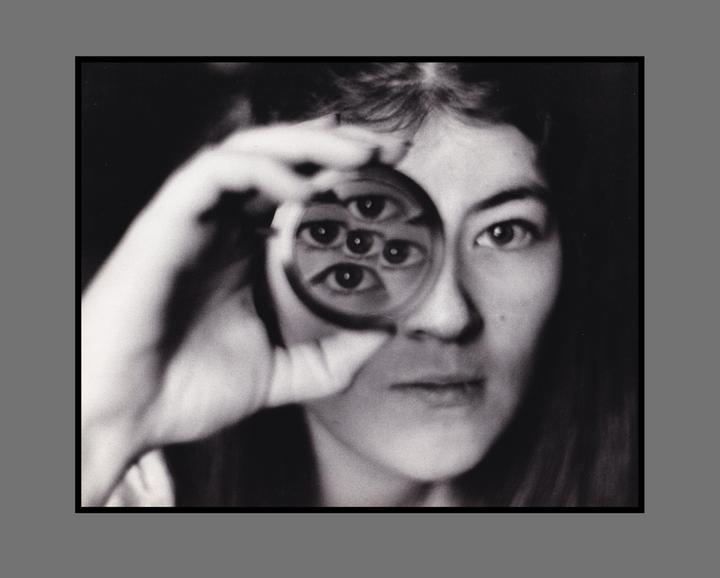 A woman holds a magnifying glass to her eye, creating a distorted reflection of multiple eyes.