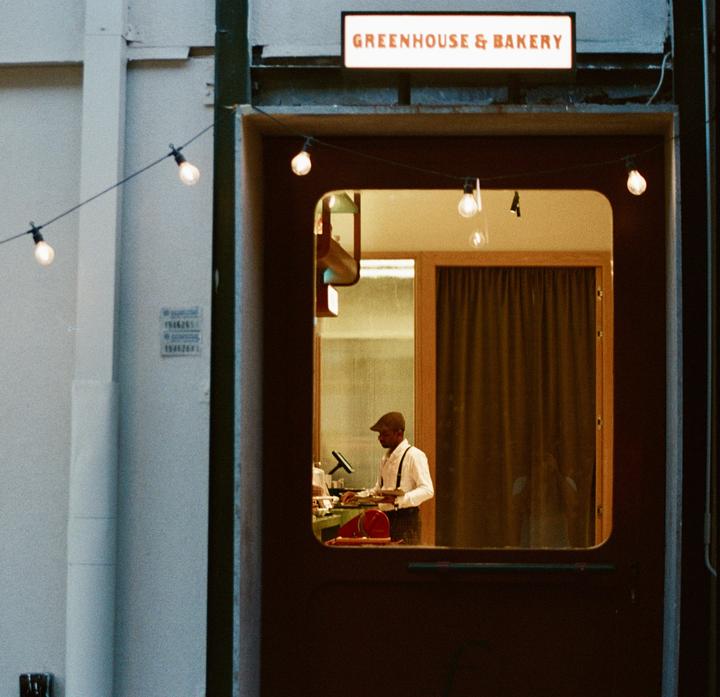 A man in a cap and suspenders works inside a bakery at night, illuminated by string lights.