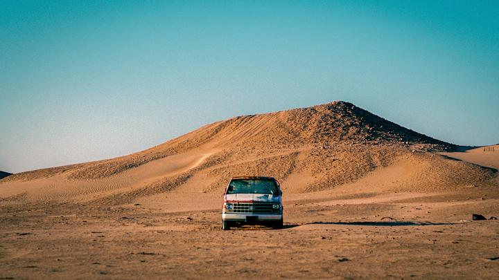 A white van is parked in a desert landscape with a large sand dune in the background.