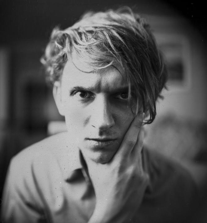 A man with messy hair rests his chin on his hand and looks directly at the viewer.