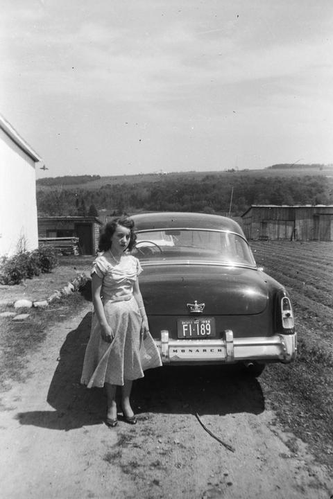 A woman in a dress stands next to a car on a dirt road with a field and hills in the background.