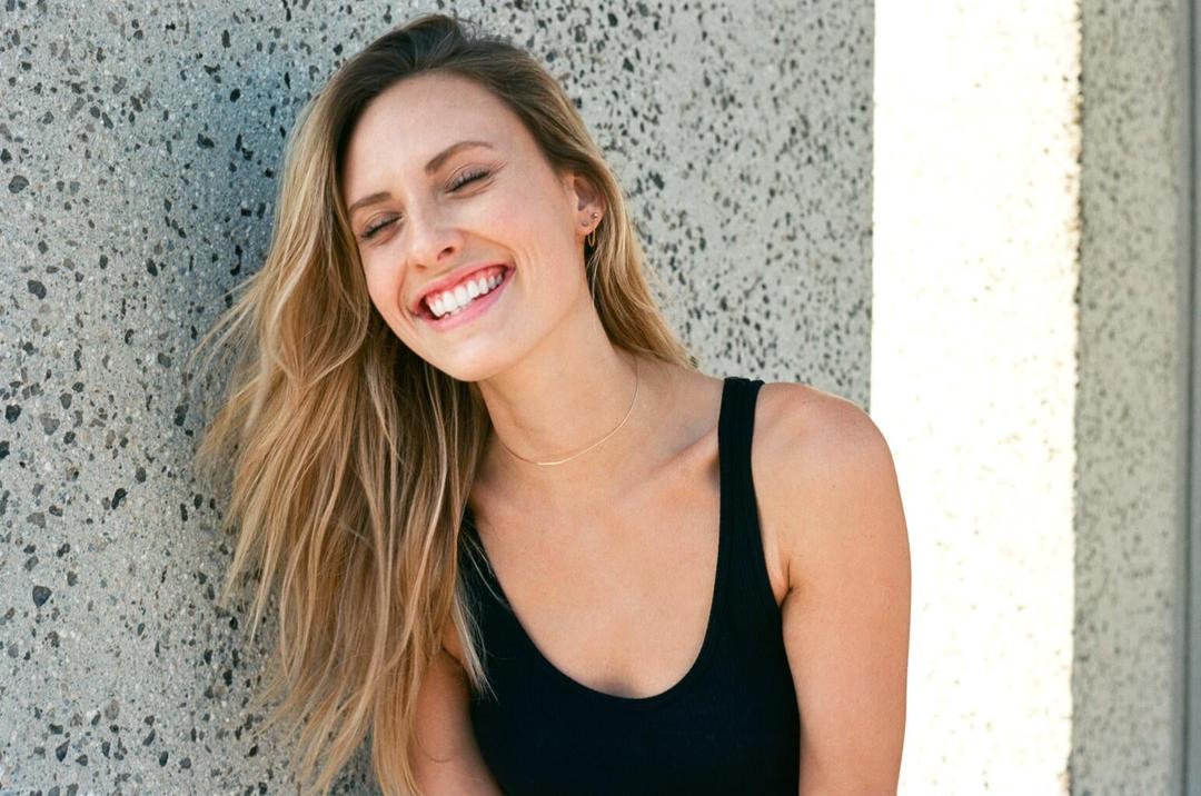 A woman with long blonde hair smiles with her eyes closed while leaning against a textured wall.