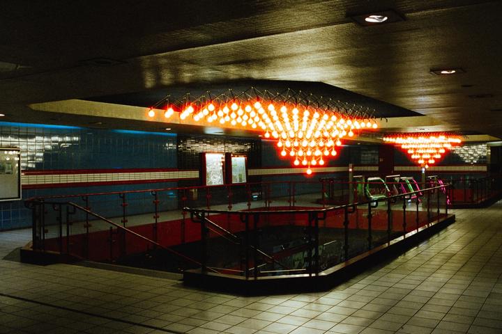 An underground station features bright orange lights arranged in a geometric pattern on the ceiling, with blue tiled walls and red accents.