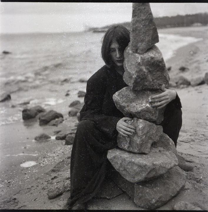 A woman sits on a rocky beach, holding a stack of balanced rocks.