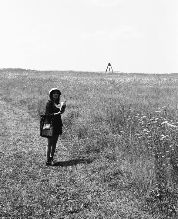 A woman wearing a hat and carrying a bag stands in a field of tall grass and wildflowers, holding a book.
