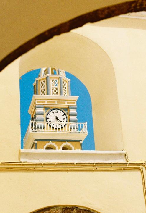 A clock tower with a white face and roman numerals is framed by a yellow archway against a bright blue sky.