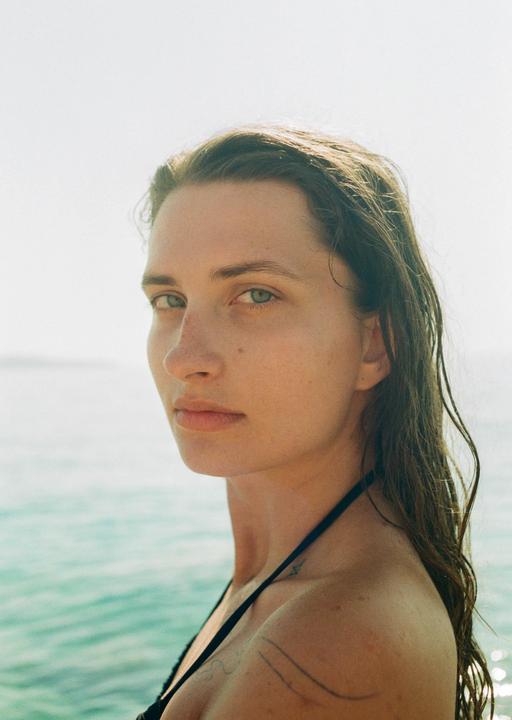 A woman with wet hair looks directly at the viewer with the ocean in the background.