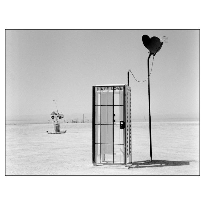 A telephone booth with a "FREE LOVE" sign stands in a desert landscape next to a pole with a heart-shaped sign.