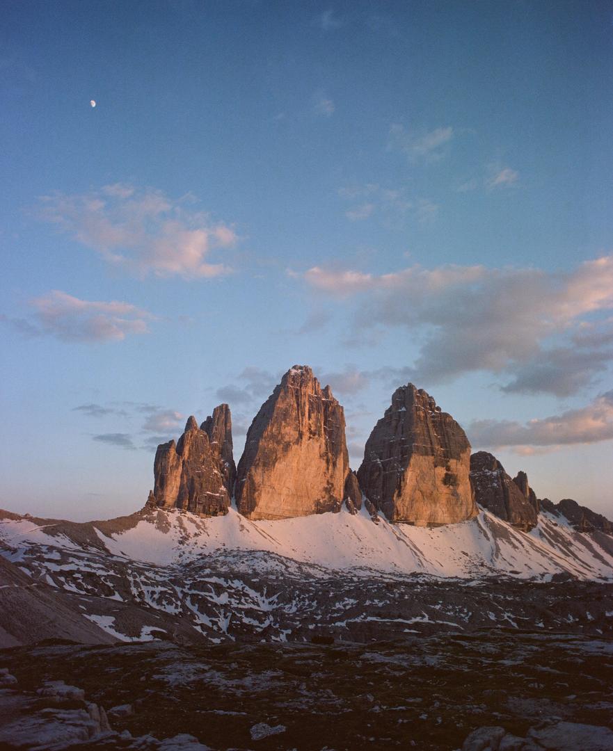 Three prominent mountain peaks are illuminated by the setting sun, with snow on their lower slopes and a crescent moon visible in the sky.