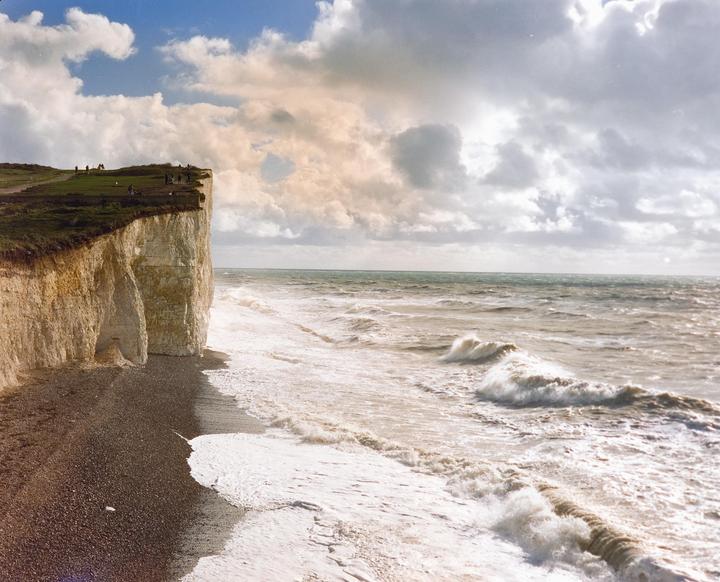 Waves crash against a rocky beach next to white cliffs with people standing on top.