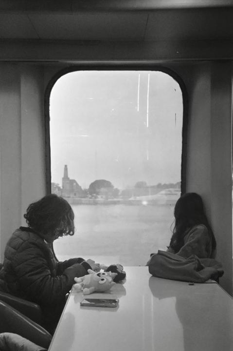Two children sit at a table looking out a window at a town across a body of water.