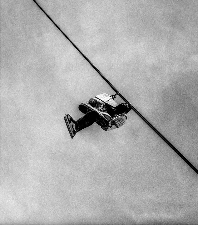 A person wearing sneakers and dark clothing is suspended in the air by a rope attached to a diagonal pole.