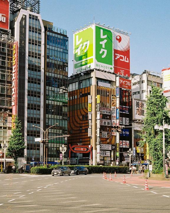 A street in a city is lined with tall buildings covered in signs and billboards, with cars driving on the road.