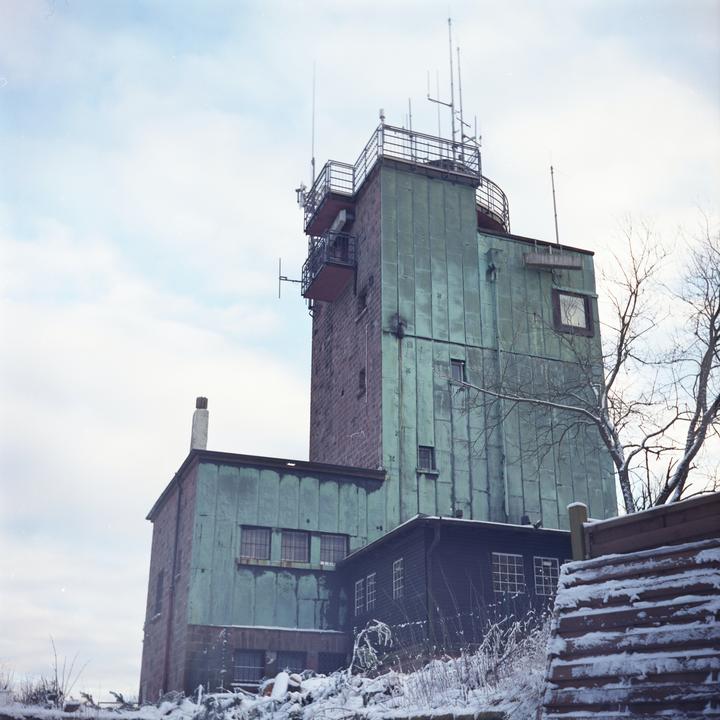 A tall building with a green facade and antennas on top stands against a cloudy sky, surrounded by snow-covered ground and bare trees.