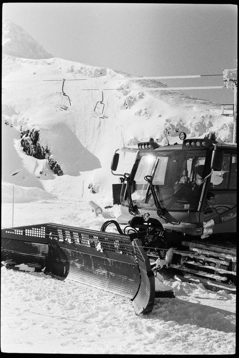 A snowcat with a large plow attachment is parked on a snowy mountain with ski lifts and mountains in the background.