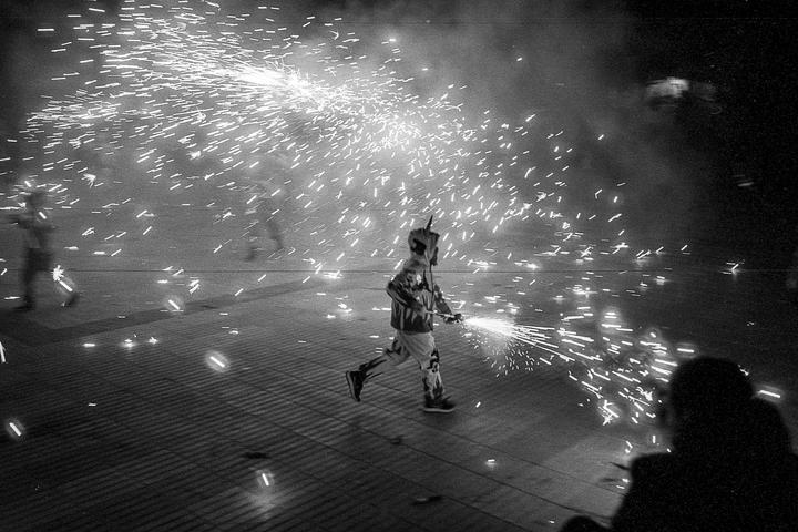 A person in protective gear runs through a street at night, holding a firework that emits sparks.