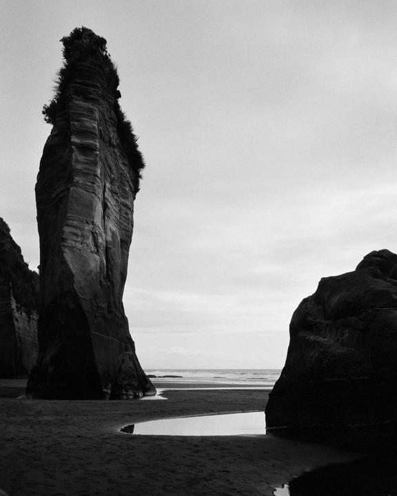 A tall rock formation stands on a black sand beach with the ocean in the background.