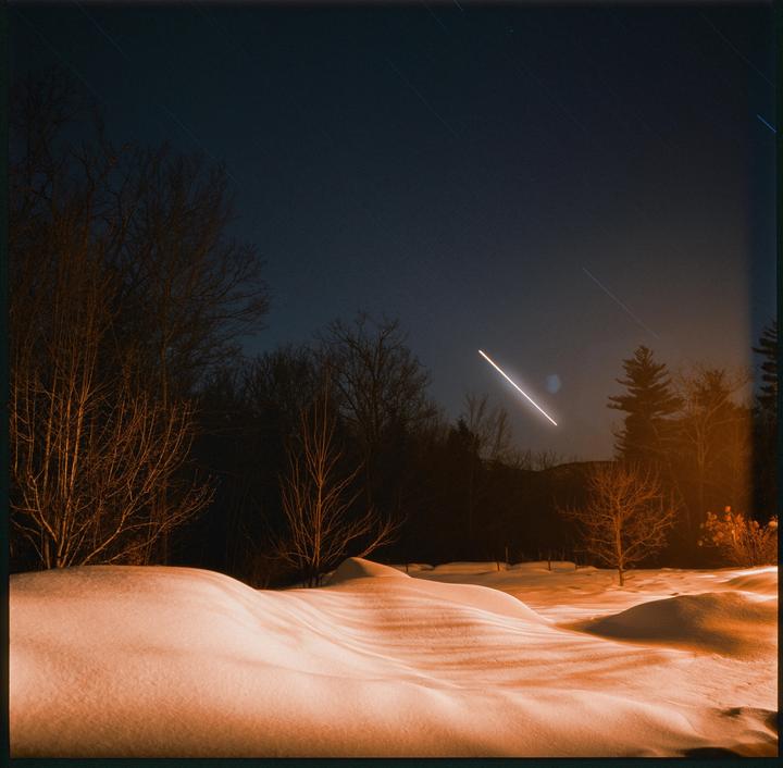 A snowy landscape at night is illuminated by an orange glow, with trees silhouetted against a dark sky showing star trails and a bright streak of light.