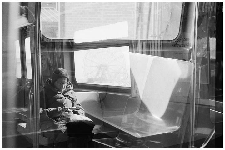A man wearing a beanie and glasses sits on a subway train, looking at his phone, with a ferris wheel visible through the window.