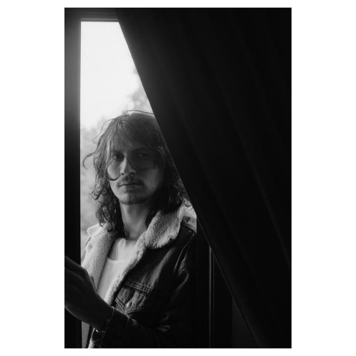 A man with long hair and a denim jacket looks out a window, partially obscured by a dark curtain.