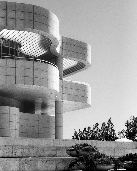 A modern building with curved walls and balconies stands above planters filled with greenery.