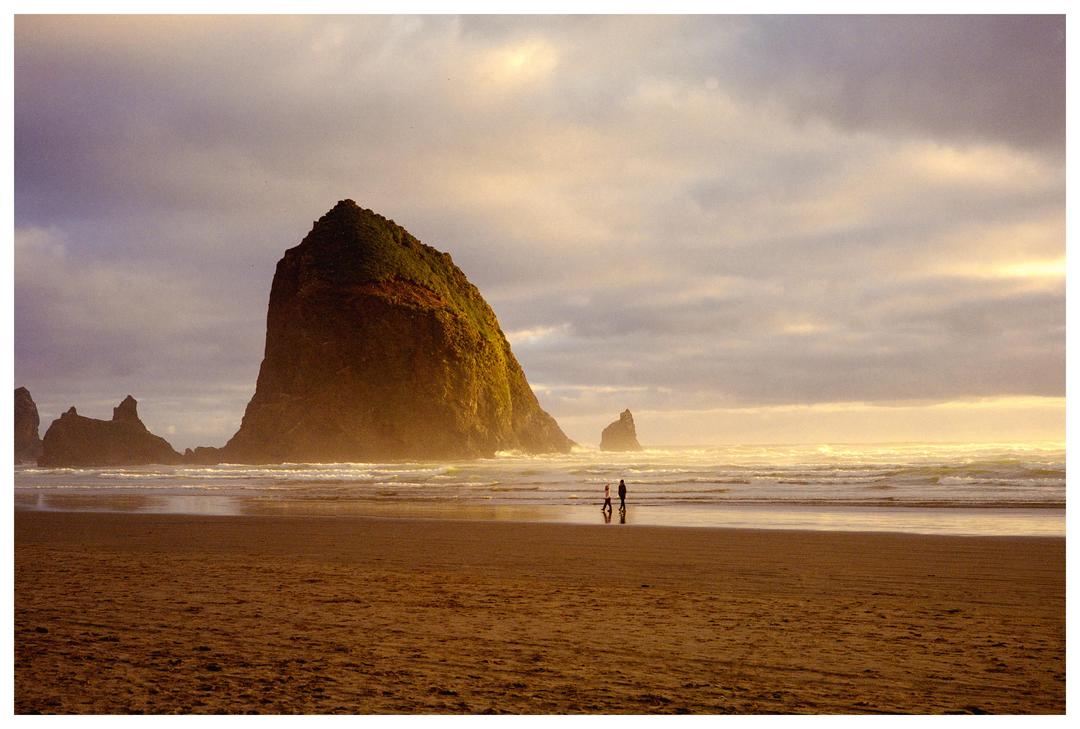 Two people walk on a beach with a large rock formation in the background under a cloudy sky.