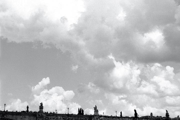 A low-angle view shows a bridge with statues and people against a cloudy sky.