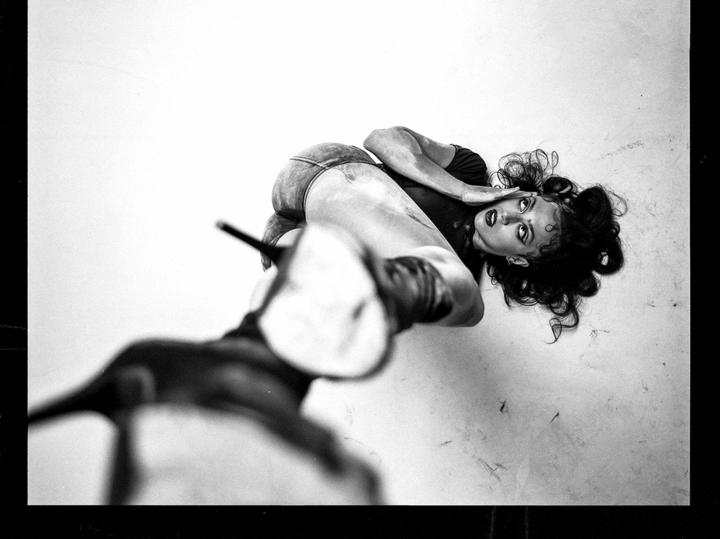 A woman with dark curly hair and dark makeup is lying on a light surface, looking up at the viewer.