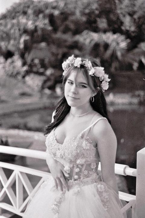 A young woman wearing a floral crown and an elaborate dress stands next to a white railing.