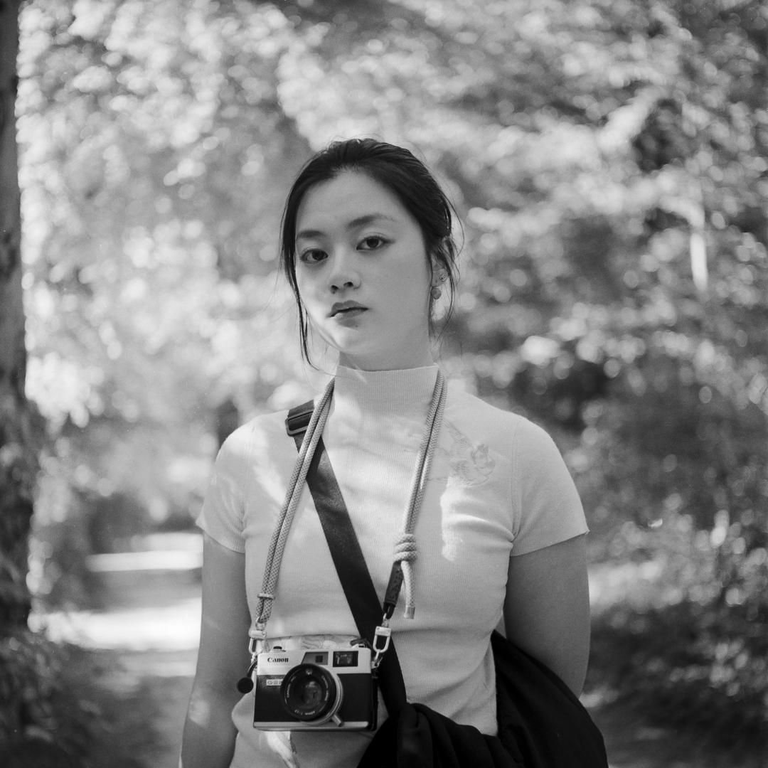 A woman wearing a turtleneck and carrying a camera stands in a forest.