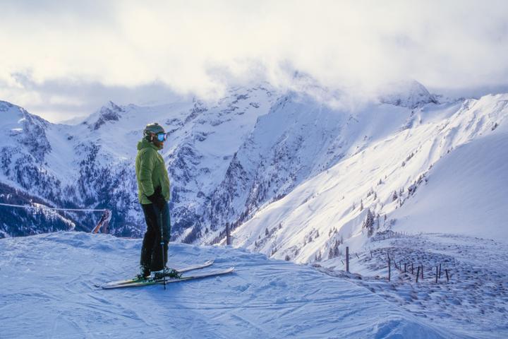 A man on skis stands on a snowy mountain looking out at a vast mountain range.