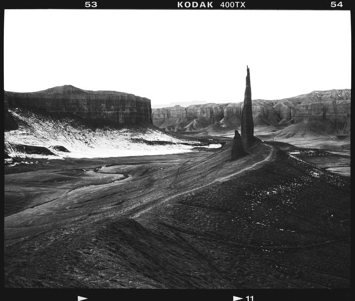A tall, thin rock formation stands in a vast, barren landscape with mesas and snow-dusted ground.
