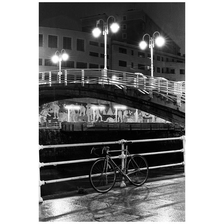 A bicycle is parked next to a railing on a wet street under a bridge at night.