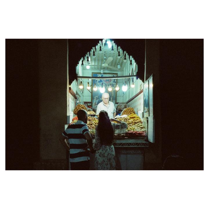 A man and a woman stand in front of a brightly lit stall filled with sweets, where a vendor stands behind the counter.