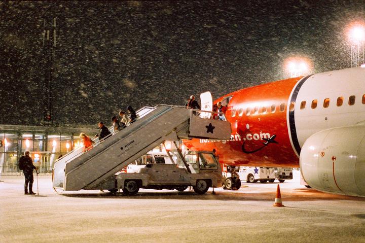People board a red and white airplane from a mobile stairway during a snowy night.