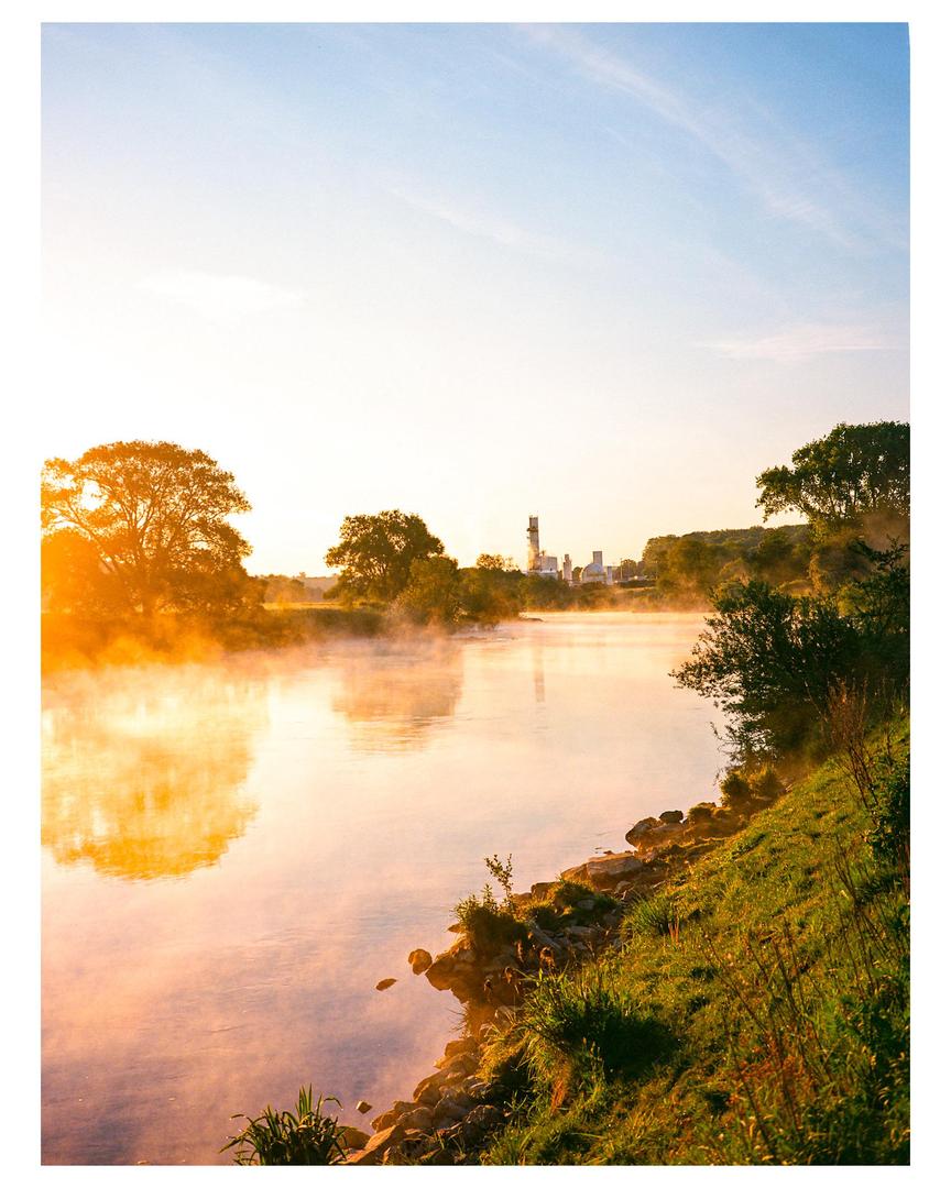 A river with mist rising from its surface flows past a grassy bank and trees under a bright sky with industrial buildings in the distance.