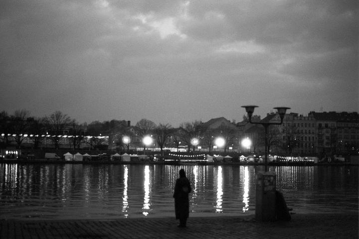 A person stands on a paved riverbank looking across the water at a city with lights reflecting on the surface at night.