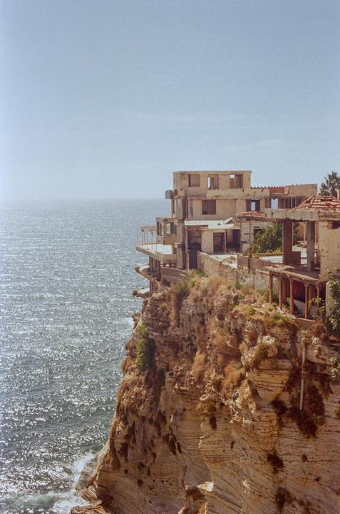An abandoned building stands on a cliff overlooking the sparkling sea under a clear sky.