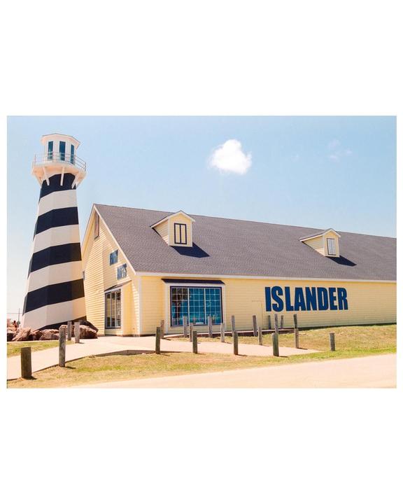 A black and white striped lighthouse stands next to a yellow building with "ISLANDER" written on its side under a blue sky with a single cloud.
