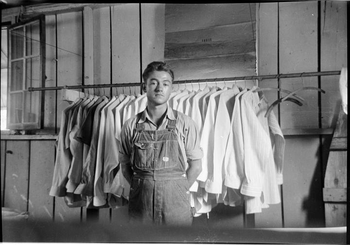 A man in overalls stands in front of a rack of shirts.