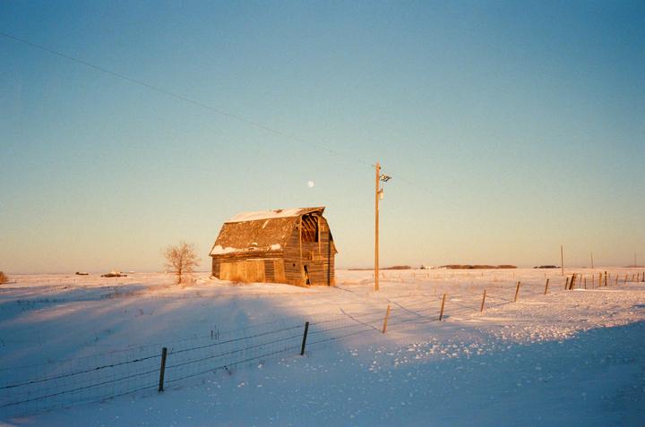 An old wooden barn stands in a snow-covered field under a clear sky with a visible moon.