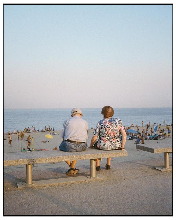 A couple sits on a bench overlooking a crowded beach with the ocean in the background.
