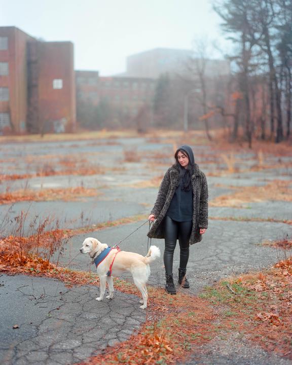 A woman walks a dog on a leash on a cracked asphalt path with autumn leaves and abandoned buildings in the background.