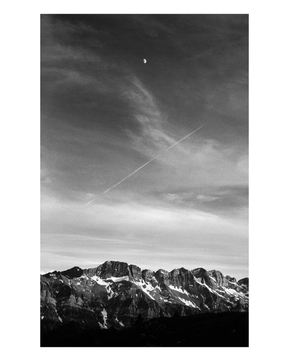 A black and white landscape shows snow-capped mountains under a cloudy sky with a moon and a contrail.